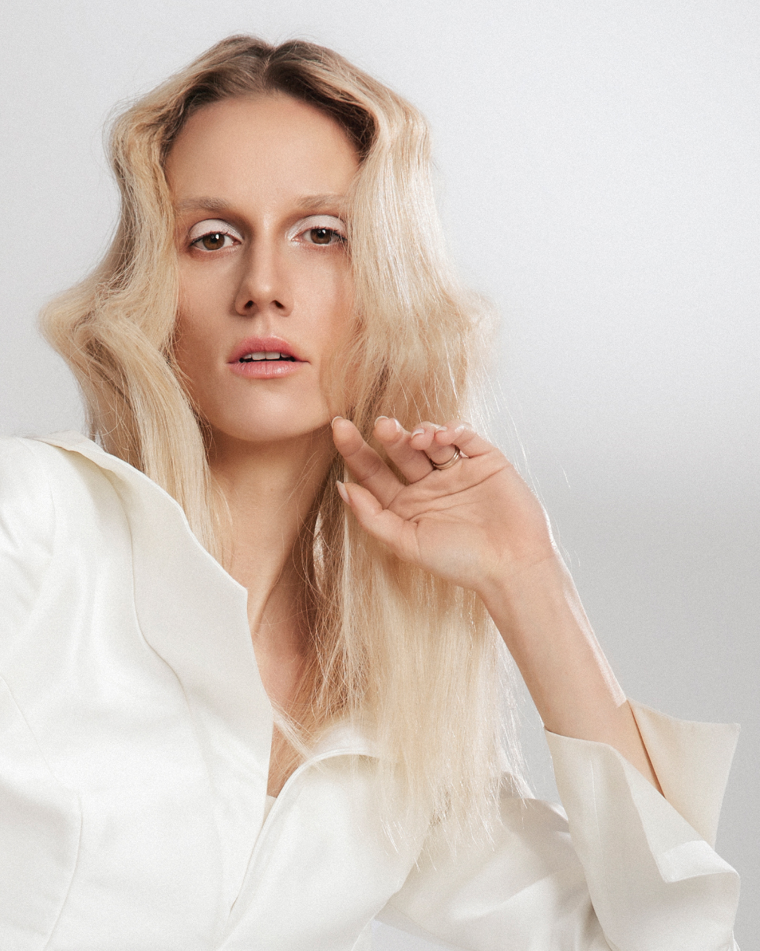 Model with soft, wavy blonde hair wearing a structured white blouse, posing elegantly with minimal makeup for a modern and sophisticated look.